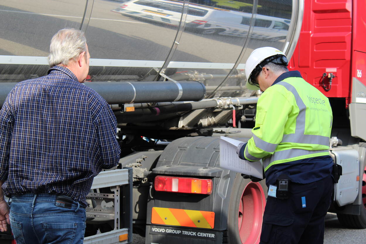 Een inspecteur staat samen met een chauffeur naast een vrachtwagen. De inspecteur bekijkt enkele papieren. In de reflectie van de vrachtwagen is een ILT-bus zichtbaar.