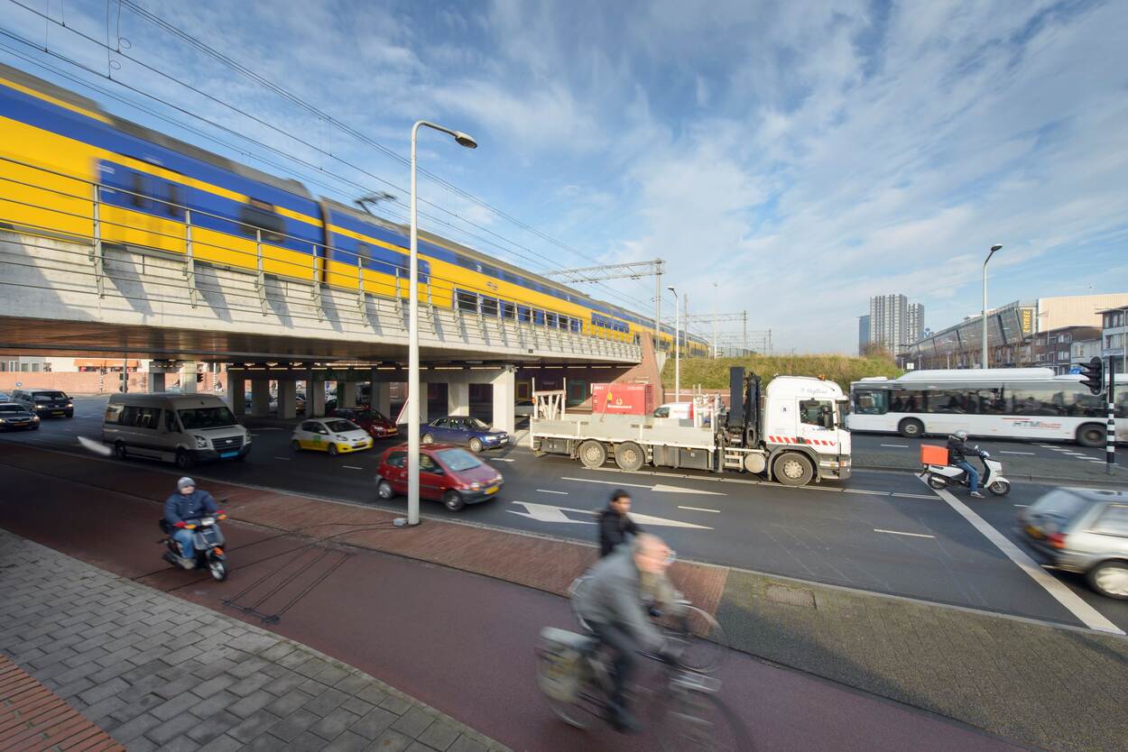 Een trein rijdt over een spoorbrug. Onder de spoorbrug rijdt een bus, een vrachtwagen, een scooter, fietsers en auto's.