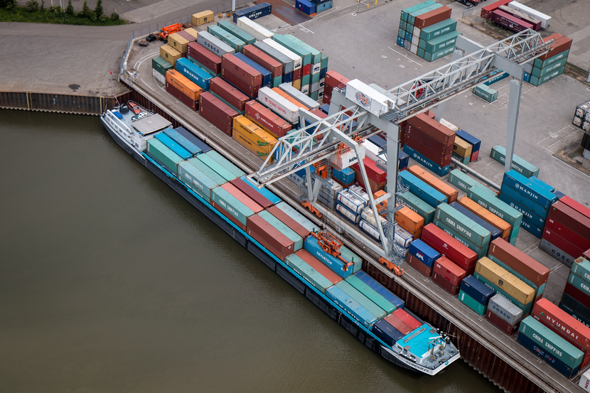 Op een haventerrein ligt een containerschip aan de kade. Containers worden in- en uitgeladen door een grote kraan.