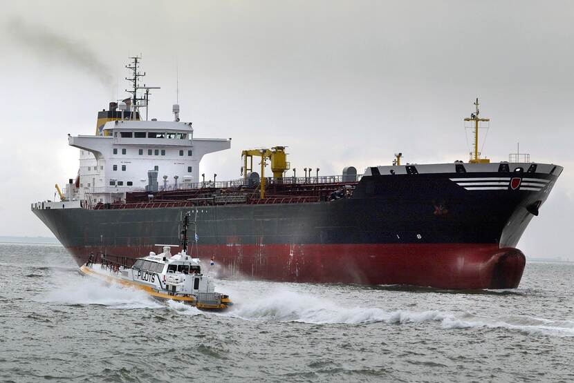 Loodsboot nadert leeg tankerschip op zee. Een donkere rookpluim van verbrande stookolie verlaat de schoorsteen van het zeeschip.