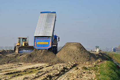 Een vrachtwagen lost een lading grond. Schuin achter de vrachtwagen staat een schovel.