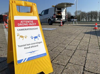Een waarschuwingsbord voor overvliegende inspectiedrones staat aan de zijkant van een parkeerplaats. Achter het bord staat een inspectie bus.