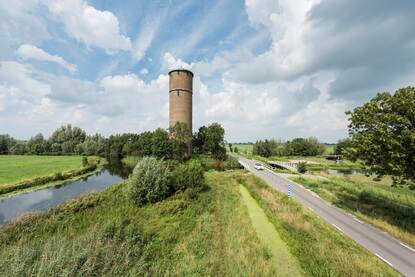 Een watertoren staat in  het landschap. Naast de watermolen loopt een klein riviertje.