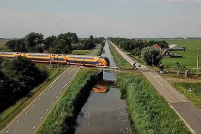 Boomrijk landschap waarin een dubbeldeks trein twee bewaakte overwegen passeert, die aan weerszijde van een sloot liggen.