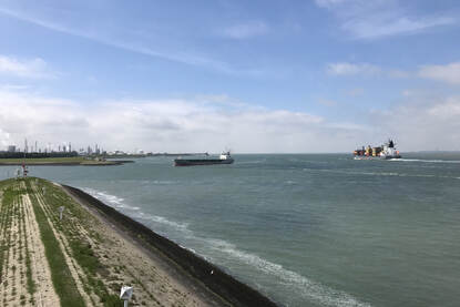 Een schip vaart een  haven in. In de verte vaart een groot zeeschip met zeecontainers.