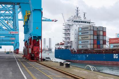 Een zeeschip met zeecontainers vaart door een havengebied.