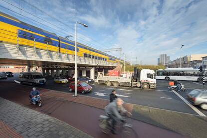 Een trein rijdt over een spoorbrug. Onder de spoorbrug rijdt een bus, een vrachtwagen, een scooter, fietsers en auto's.