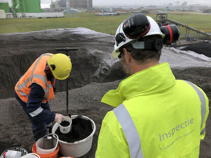 Een inspecteur kijkt toe hoe een monster wordt genomen van een berg termisch gereinigde grond.