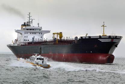 Loodsboot nadert leeg tankerschip op zee. Een donkere rookpluim van verbrande stookolie verlaat de schoorsteen van het zeeschip.