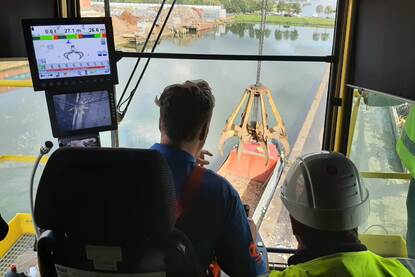 Foto vanuit cabine van een kraan met grote stalen grijper die afval tussen kade en schip overbrengt.